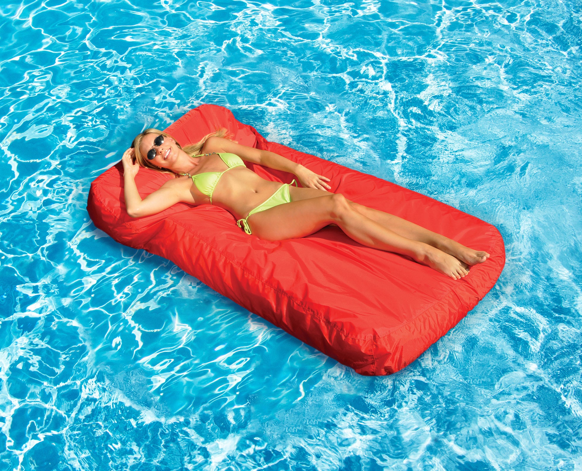 Woman in a green bikini lying on a red inflatable raft in a pool.