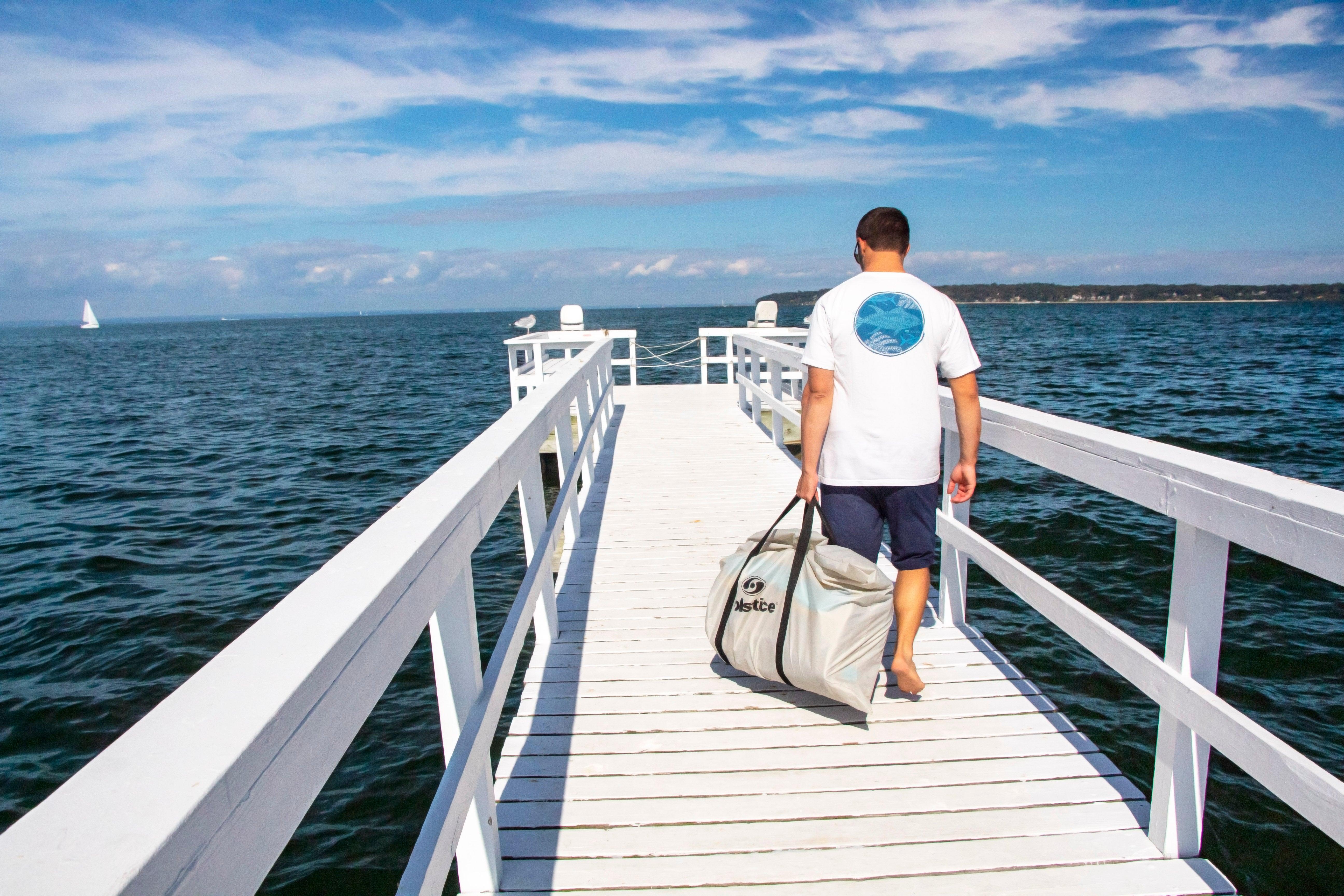 Man walking barefoot on white wooden pier carrying large bag with ocean and sailboat in background