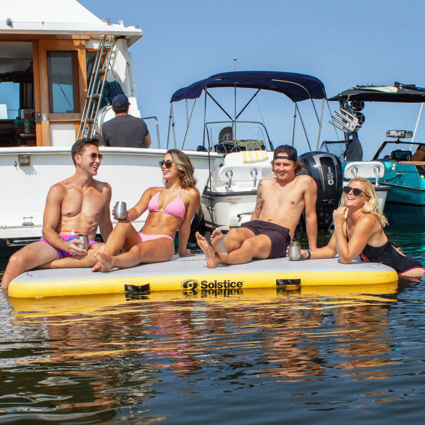 Four people relaxing on a yellow inflatable platform in the water with boats in the background.