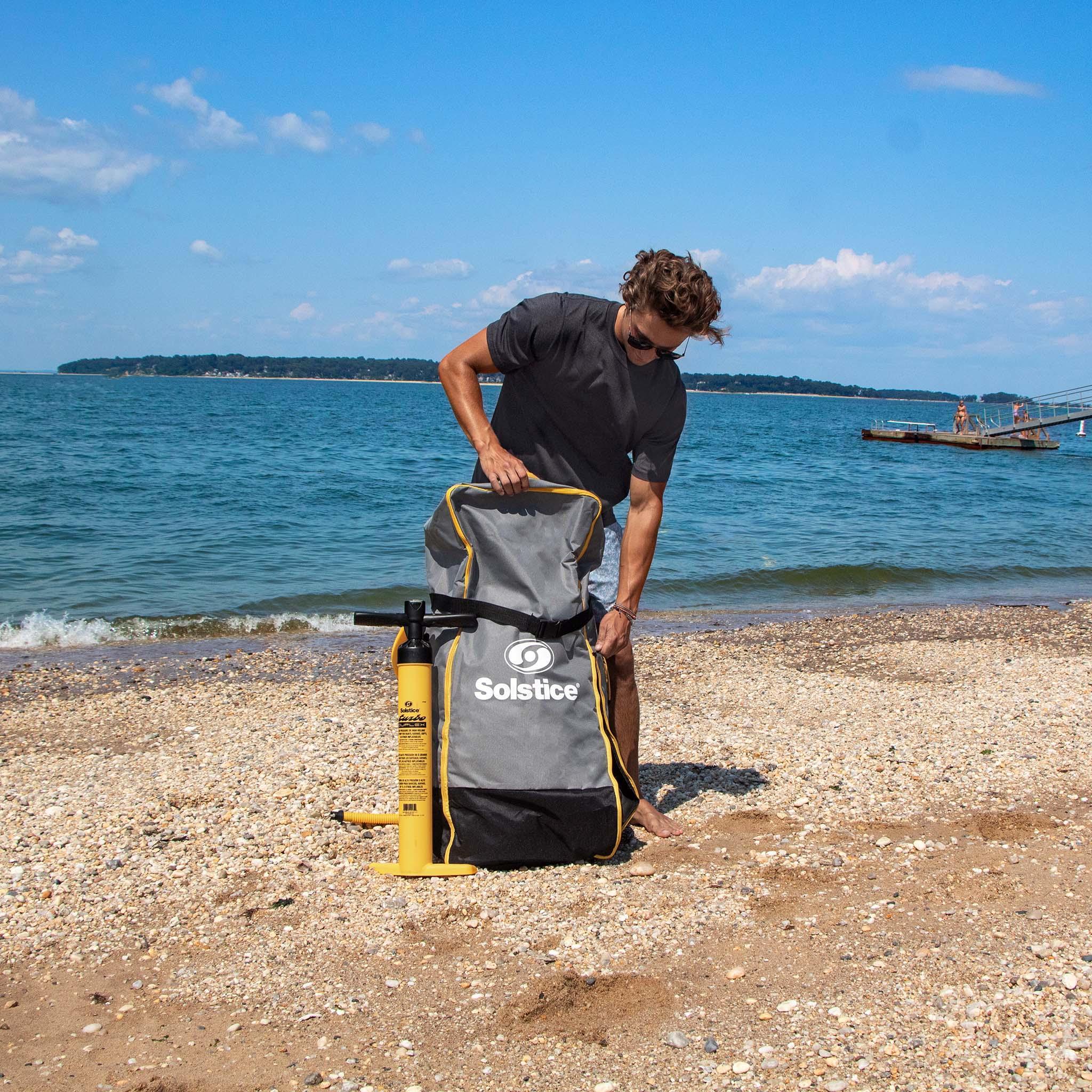 Man on beach opening large gray Solstice touring backpack with yellow pump beside him