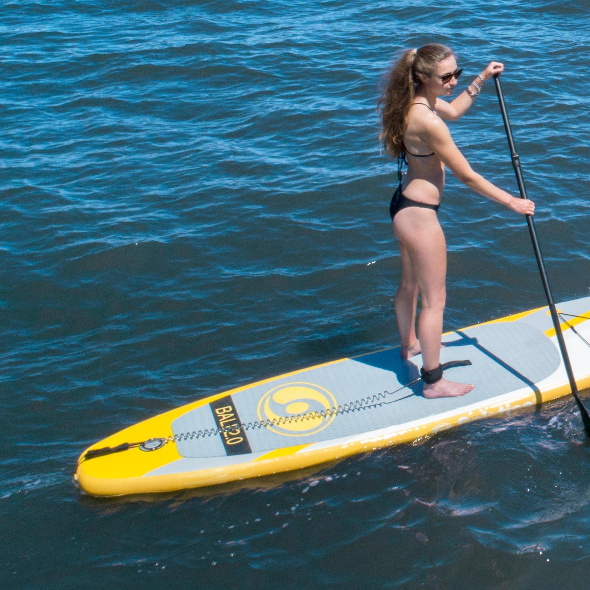 Woman paddleboarding on blue water using yellow and gray BAL2.0 stand-up paddleboard with paddle leash